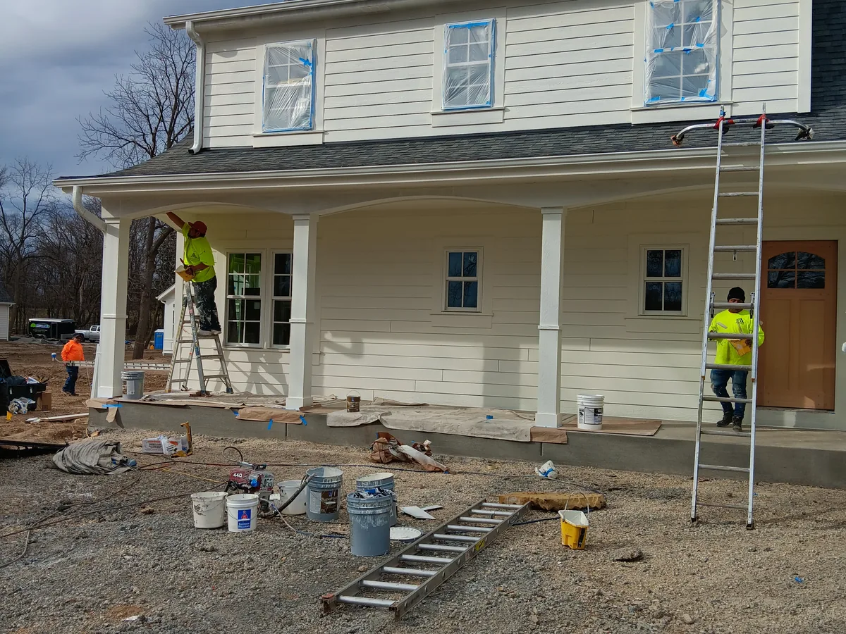 Painters on ladders working on the front porch and siding of a white farmhouse