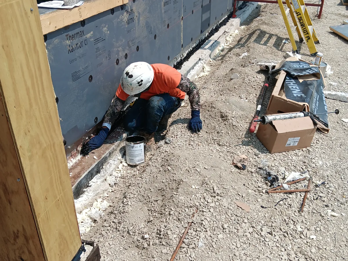 Worker in a hard hat applying waterproofing at the base of a foundation wall