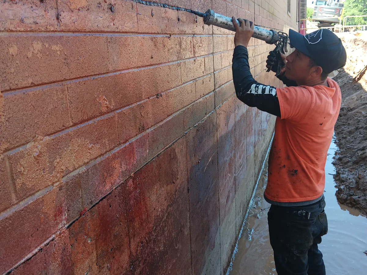 Worker applying liquid waterproofing to a poured concrete wall with a hand applicator
